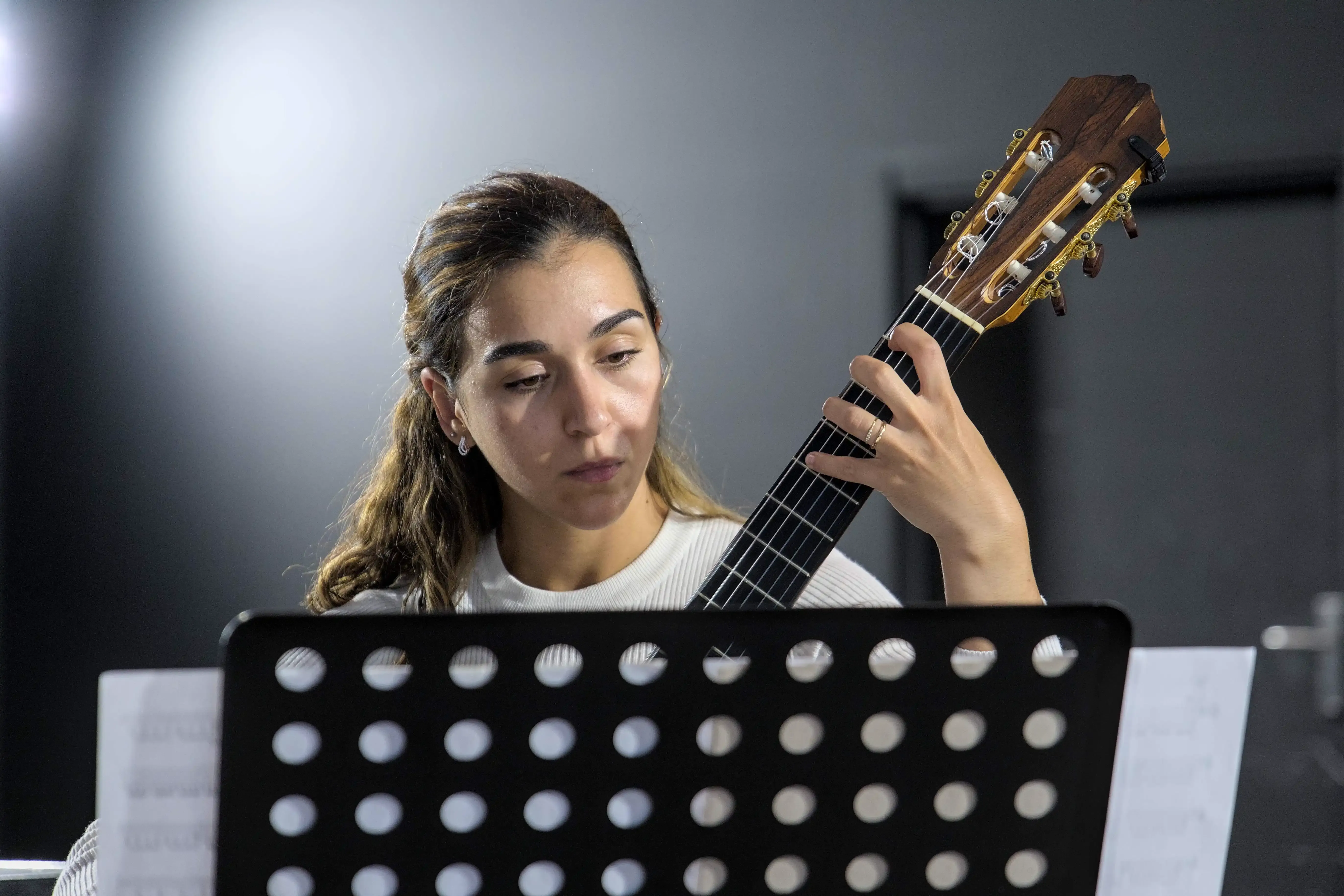 Uma mulher toca guitarra clássica enquanto olha a partitura. Ela tem cabelo claro apanhado e tem vestida uma camisola branca.